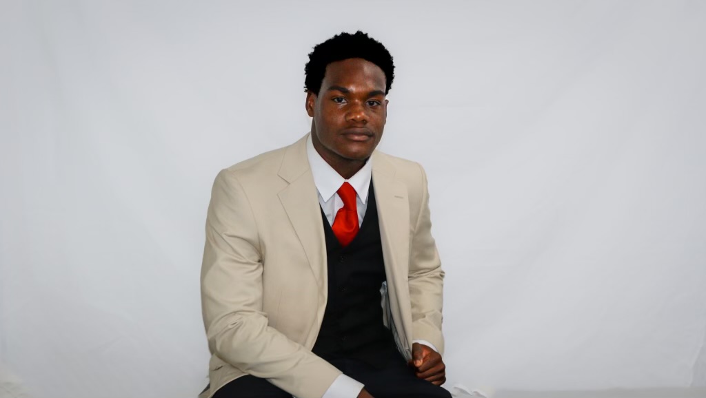 AJ seated in a tan suit with a red tie