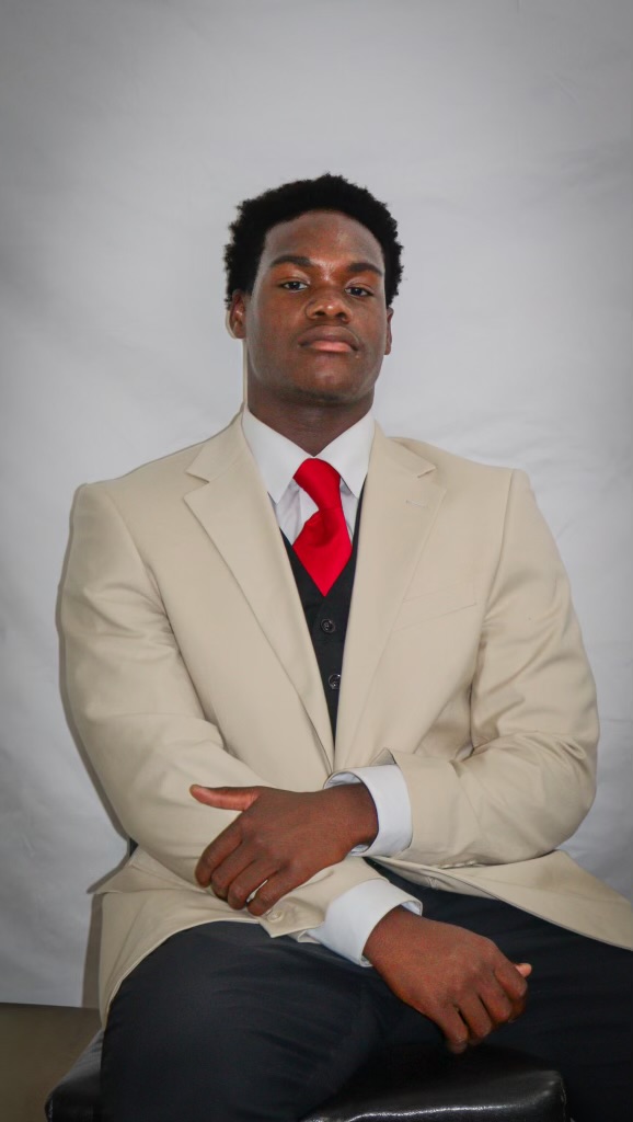 AJ seated with hands folded, dressed in a tan suit